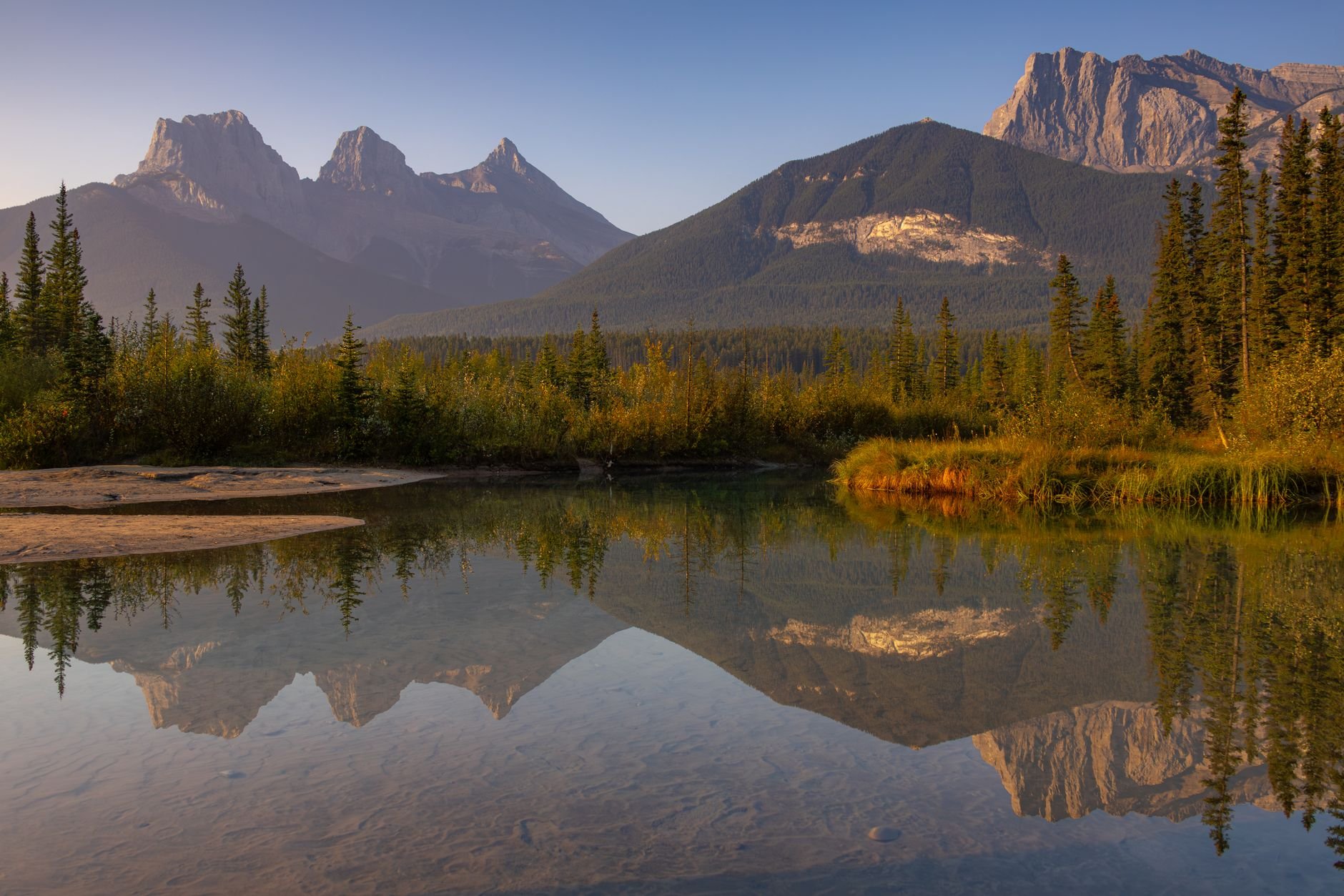 Golden hour reflection of the Three Sisters peaks in Canmore, Alberta, creating a peaceful landscape.